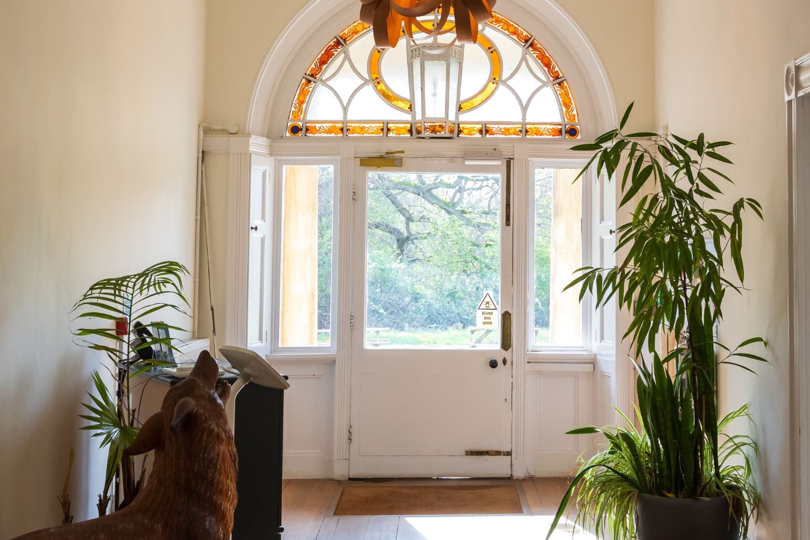Bright foyer with white door and amber stained-glass arched transom, tall potted plants flanking the entrance and wooden animal sculpture.