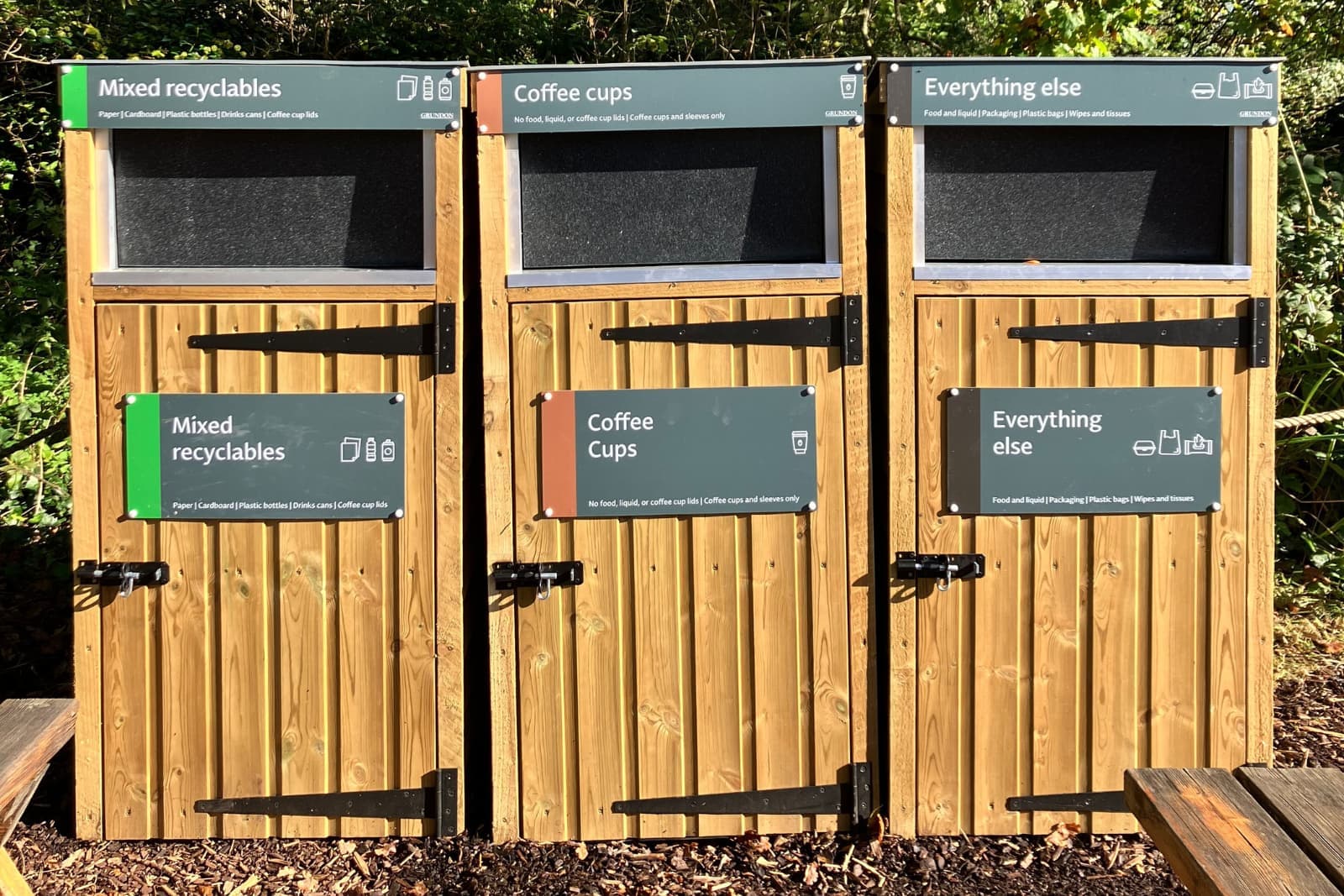 Three wooden outdoor bins with green signs labeled "Mixed recyclables", "Coffee Cups", and "Everything else."