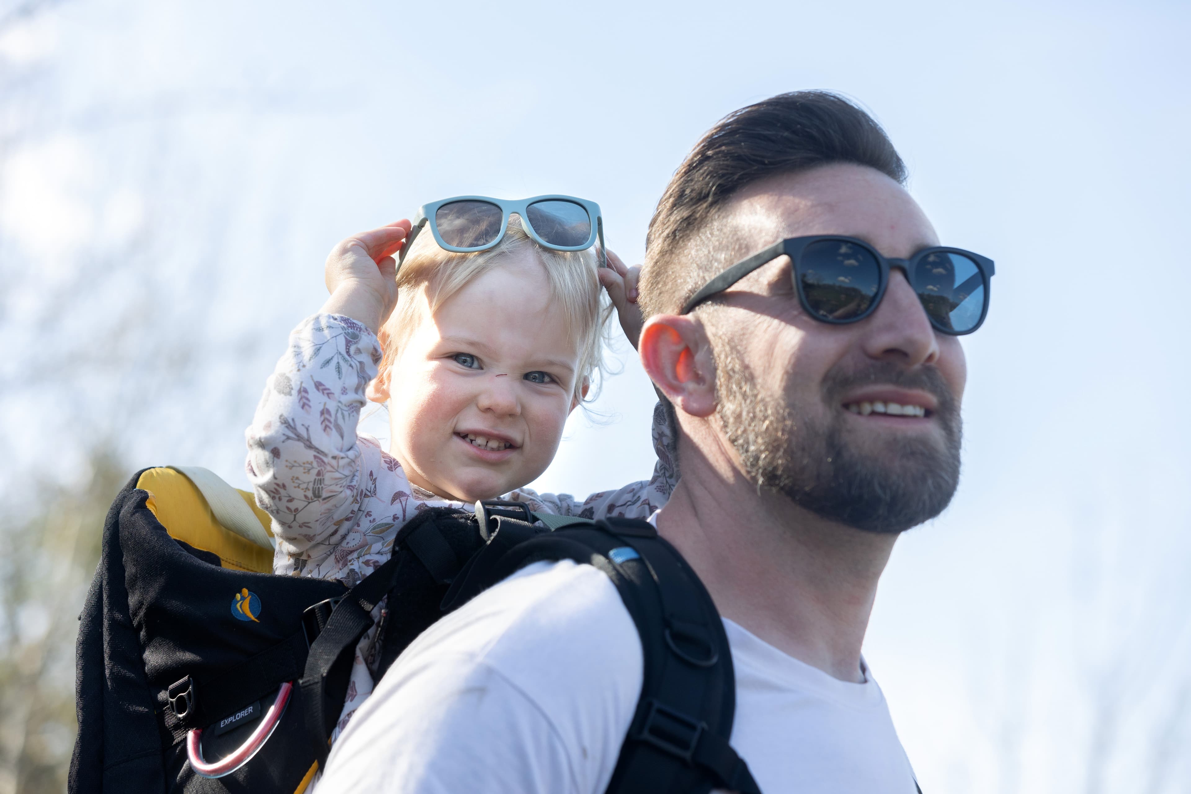 A dad has his daughter on his back in a carrier. She is smiling at the camera whilst raising sunglasses of her head.