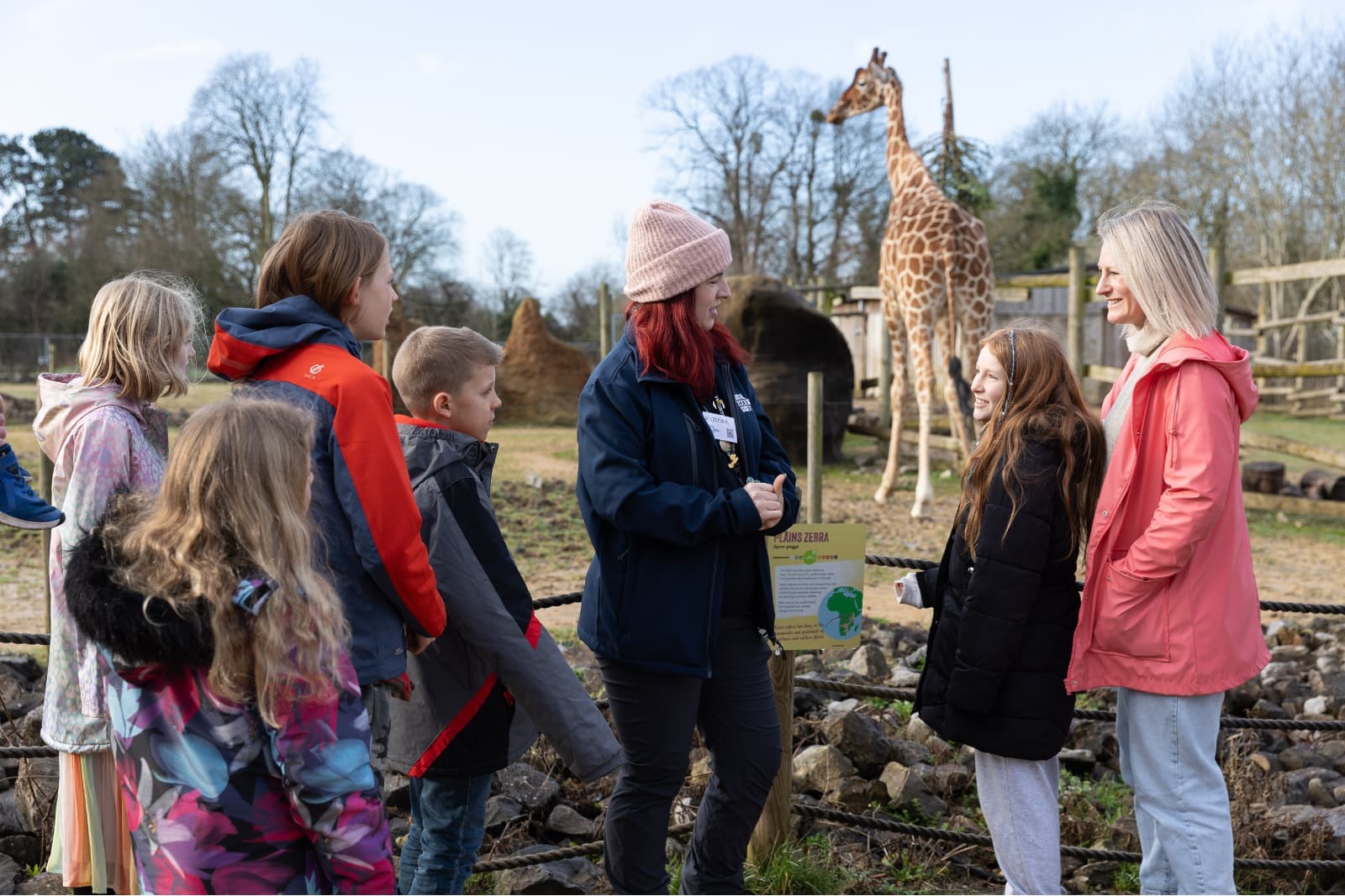 Zoo guide talks to children and an adult beside a giraffe enclosure as the giraffe stands in the background.