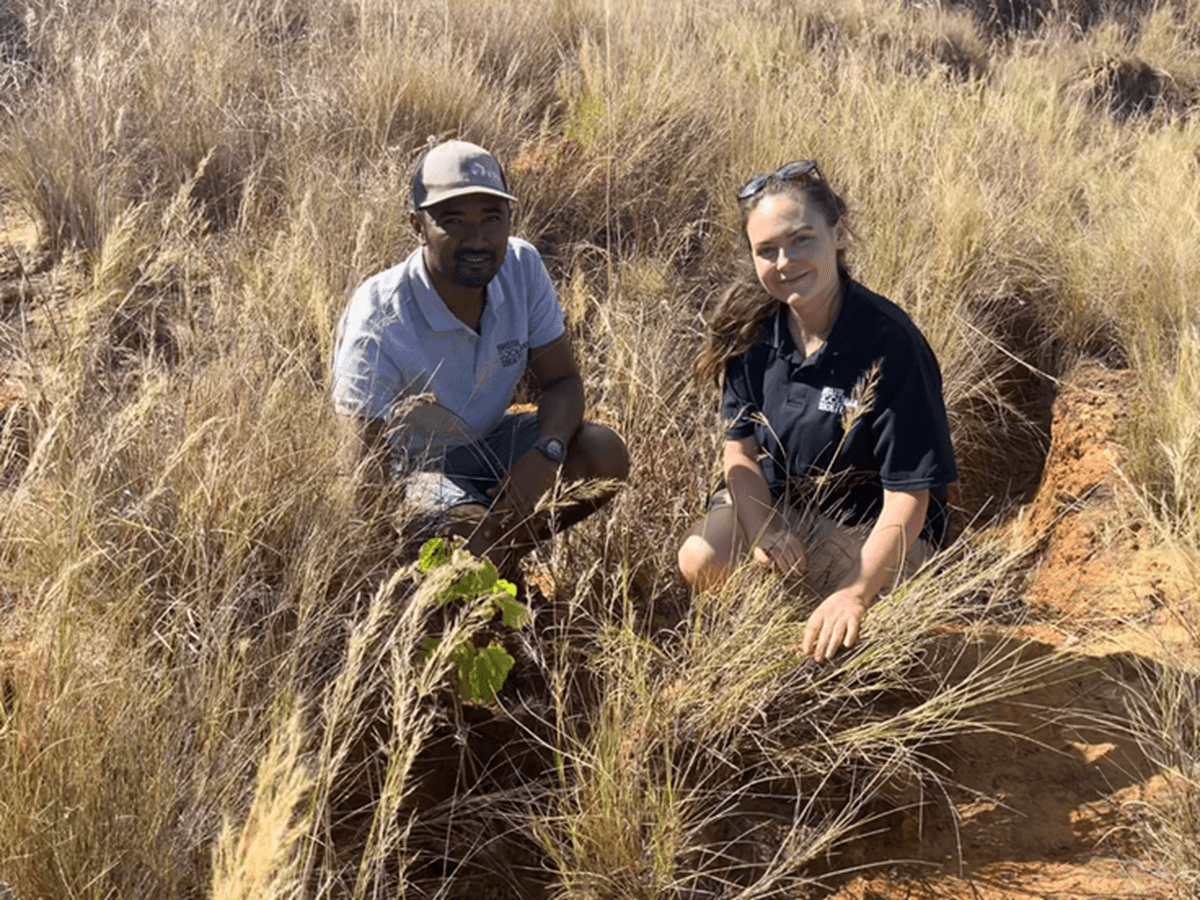Two people kneel in a grassy field, smiling at the camera. A small plant grows in front of them, surrounded by dry grass.